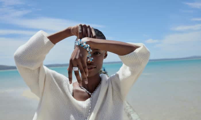 Mujer con jersey blanco mostrando pulsera y collar Mikado de Tamara Comolli frente a un mar turquesa